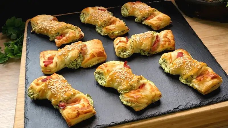 Goldbraun gebackene, gedrehte Blätterteig-Stangen mit Bärlauchcreme, knusprigem Speck und Sesam auf einem rustikalen Holzbrett