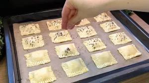 Vorbereitete Blätterteigtaschen auf dem Backblech, die mit Sesam bestreut werden.