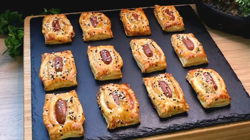 Goldbraun gebackene Käse-Würstchen-Häppchen aus Blätterteig mit Sesambestäubung auf einem Teller