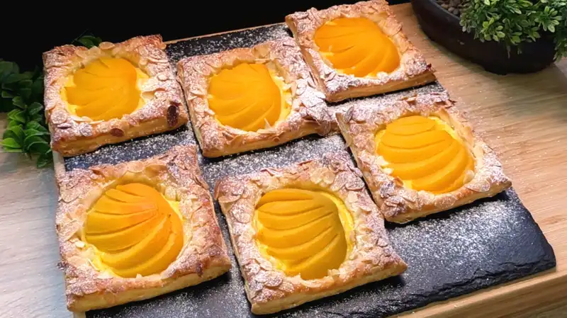Goldbraun gebackene Mini-Pies aus Blätterteig mit Vanillepudding, Pfirsichhälften und Mandelblättchen.