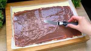 Ein Löffel verteilt dunklen Schokoladenpudding auf der hellen Blätterteigplatte