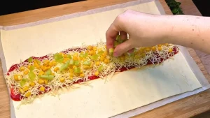 Die Zutaten (Sauce, Schinken, Mais, Paprika) werden auf dem mittleren Streifen des Teigs verteilt