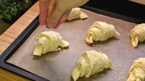 Eine Hand streut Sesamsamen auf die mit Ei bestrichenen, rohen Croissants