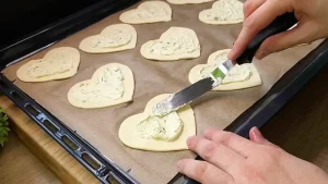 Kräuterfrischkäse wird gleichmäßig auf die ausgestochenen Blätterteig-Herzen gestrichen.