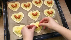 Zwei Mini-Würstchen werden auf das mit Käse bestreute Blätterteig-Herz gelegt.
