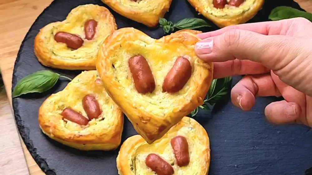 Detailansicht eines Blätterteig-Herzens mit geschmolzenem Käse und zwei Würstchen.