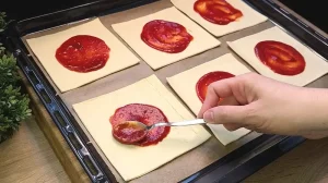 Tomaten-Pizzasoße wird gleichmäßig auf die ausgeschnittenen Blätterteig-Quadrate gestrichen