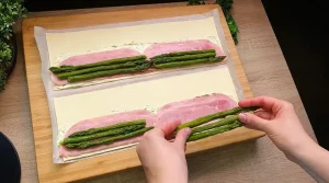 Grüner Spargel wird auf den mit Kräuterfrischkäse und Schinken belegten Blätterteig platziert.