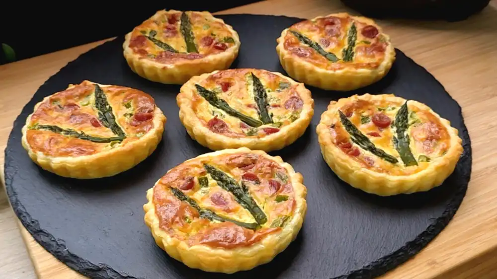 Sechs goldbraun gebackene Mini-Quiches mit grünem Spargel und Salami in Förmchen.