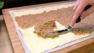 Eine dunkle Walnussfüllung wird gleichmäßig auf eine frische Blätterteigplatte gestrichen.
