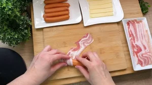 Ein Wiener Würstchen wird mit Käse gefüllt und fest in eine Scheibe Speck eingerollt