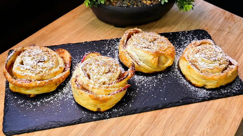 Wunderschön geformte, goldbraun gebackene Blätterteig-Apfelrosen mit einer Kruste aus gemahlenen Walnüssen, fein mit Puderzucker bestäubt auf einer edlen Kaffeetafel.