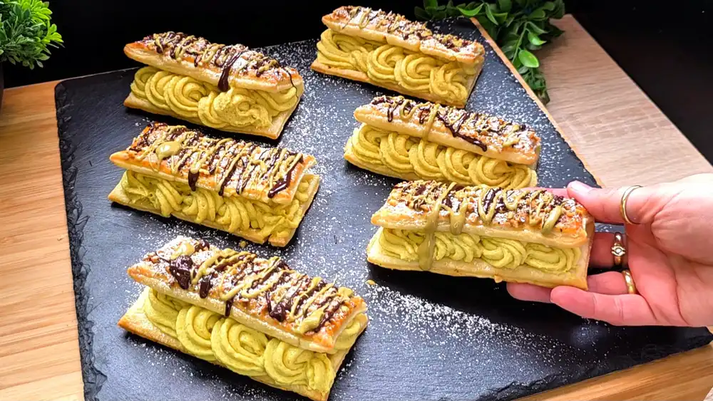 Goldbraun gebackene Blätterteig-Schnittchen, gefüllt mit Pistaziencreme, verziert mit geschmolzener Schokolade, Pistazienmus und Puderzucker.