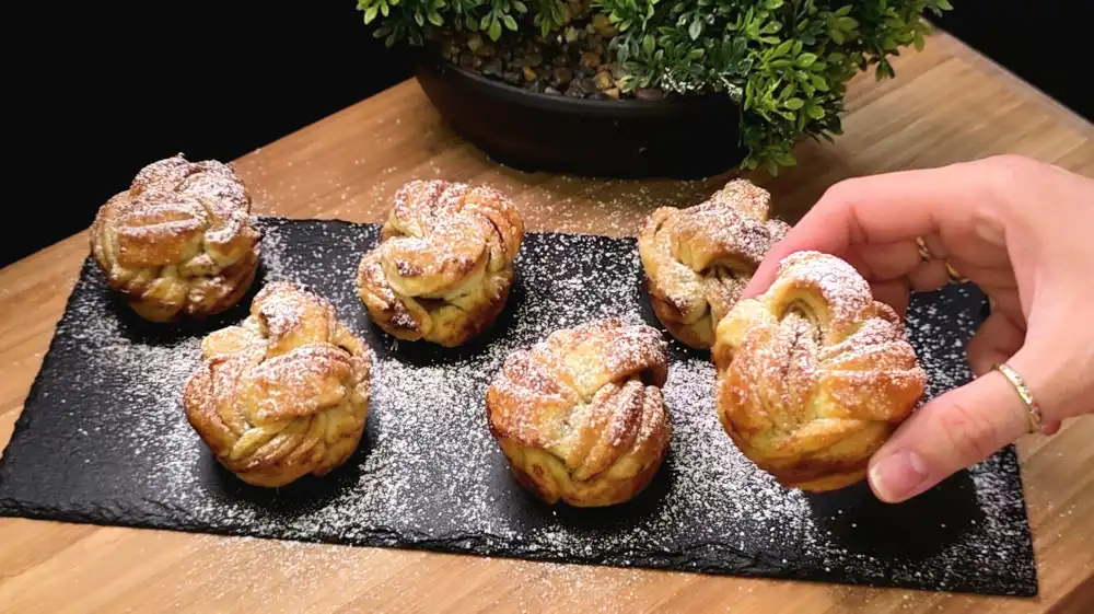 Goldbraun gebackene, geflochtene Blätterteig-Zimtknoten mit Puderzucker in einer Muffinform.