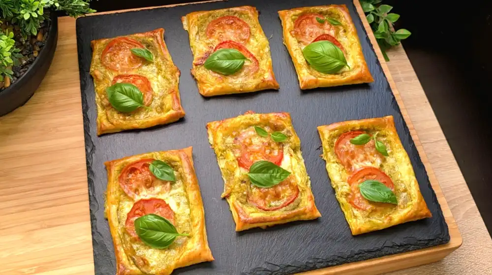Goldbraun gebackene Blätterteig-Schnittchen belegt mit grünem Pesto, Tomatenscheiben, geschmolzenem Mozzarella und frischem Basilikum.