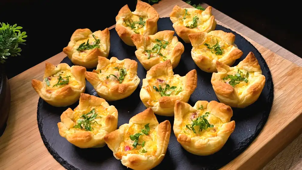 Zwölf goldbraun gebackene, krosse Blätterteig-Eiermuffins mit geschmolzenem Käse, Paprikawürfeln und Frühlingszwiebeln auf einem rustikalen Holzbrett.