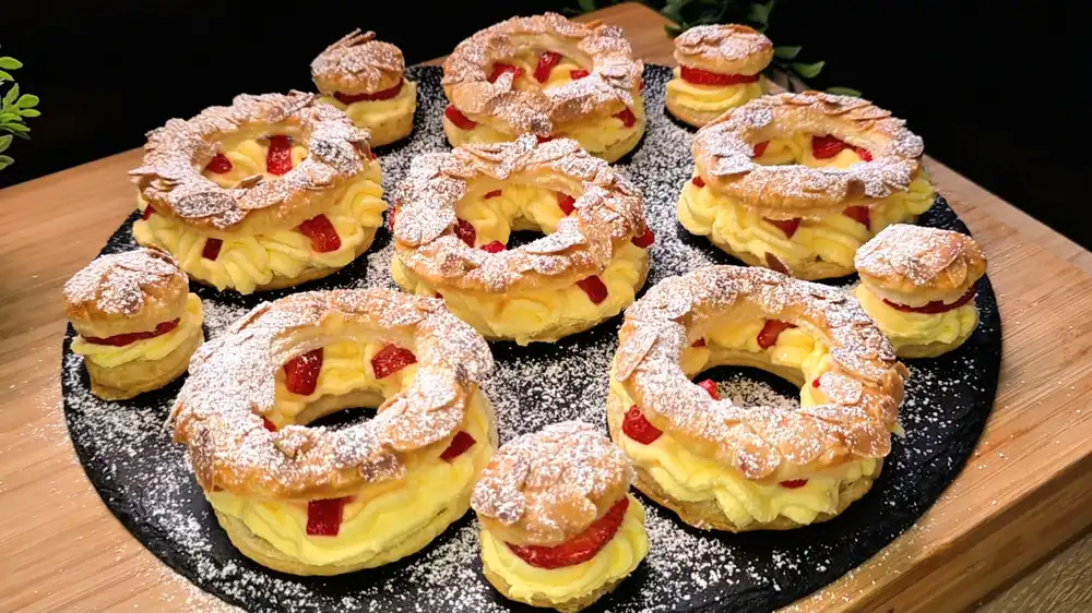 Goldbraun gebackene Blätterteigringe mit gehobelten Mandeln, gefüllt mit aufgespritzter Vanillecreme und frischen Erdbeerscheiben, bestäubt mit Puderzucker.
