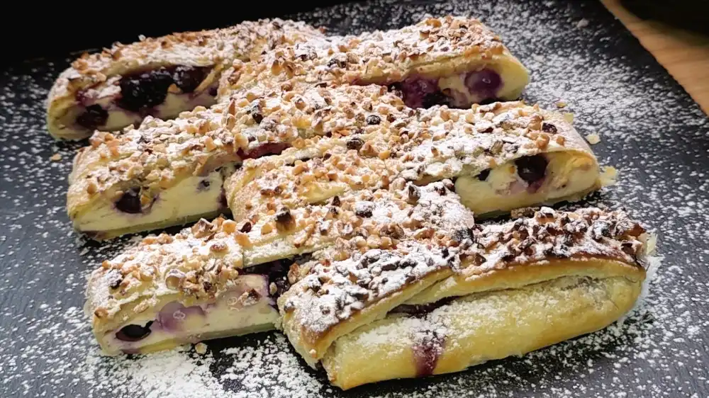 Ein goldbraun gebackener Blätterteig-Quark-Strudel mit Heidelbeeren auf einem Kuchengitter, üppig mit gehackten Haselnüssen und Puderzucker bestäubt.