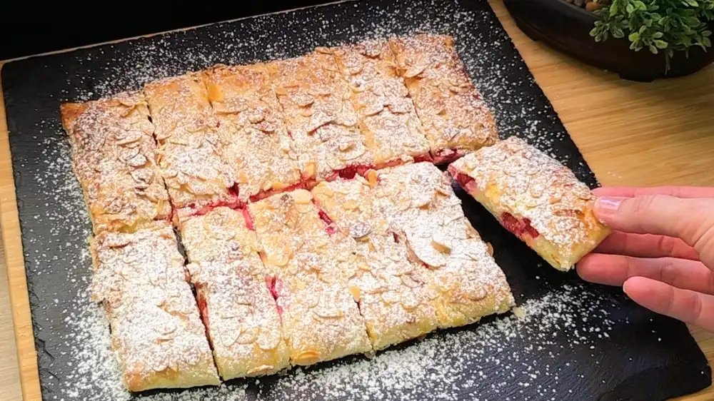 Goldbraun gebackene Blätterteig-Riegel mit gehobelten Mandeln und Puderzucker, gefüllt mit Quarkcreme und roten Johannisbeeren auf einem sommerlichen Holztisch.