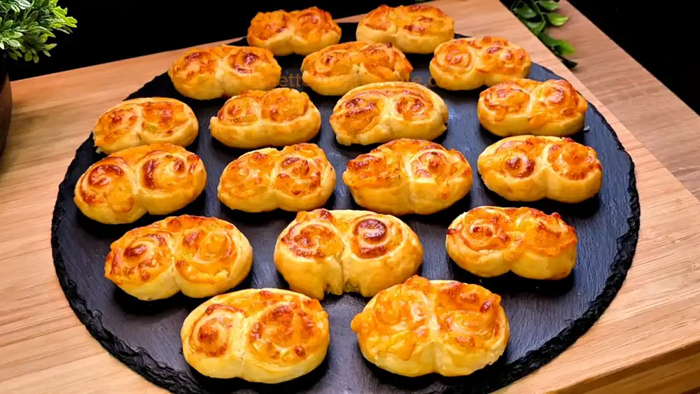 Goldbraun gebackene herzhafte Blätterteig-Öhrchen mit einer sichtbaren Füllung aus Käse, Chili-Quark und grüner Paprika auf einem rustikalen Holzbrett.