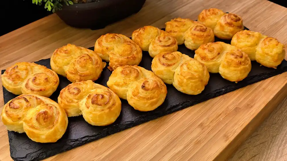 Eine Etagere voller goldbraun gebackener, krosser Blätterteig-Käse-Öhrchen mit Kräuterfrischkäse auf einer sommerlichen Kaffeetafel im Garten.
