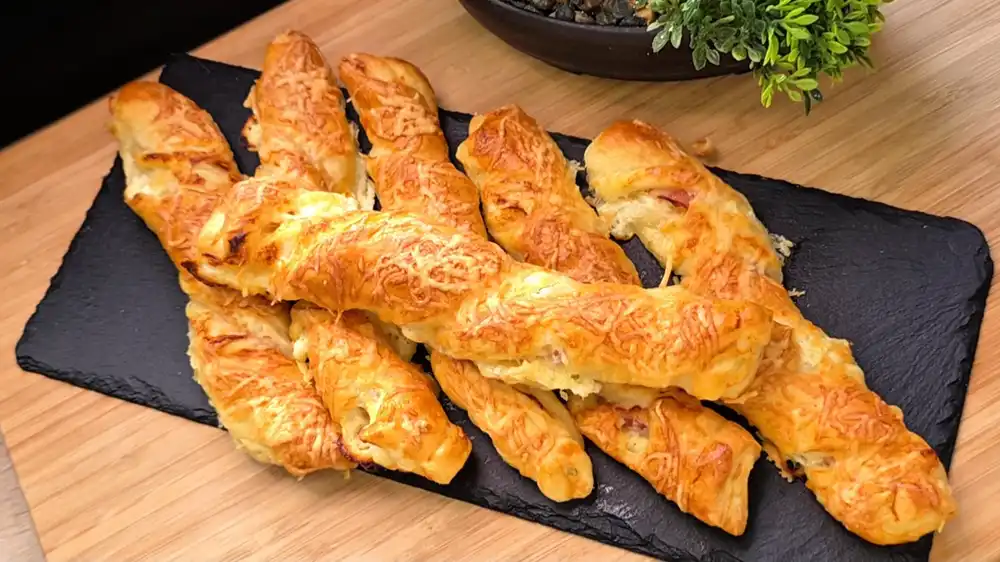 Sechs goldbraun gebackene, gedrehte Blätterteig-Käse-Speck-Stangen mit einer krossen Käsekruste auf einem rustikalen Holzbrett.
