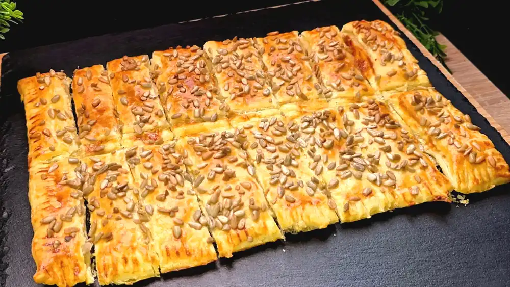 Goldbraun gebackene Blätterteig-Käsestangen mit einer Kruste aus gerösteten Sonnenblumenkernen auf einem rustikalen Holzbrett.