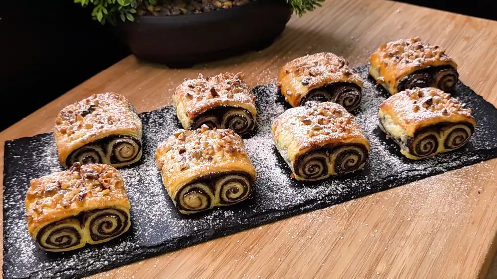 Eine Servierplatte voller goldbraun gebackener, krosser Blätterteig-Schoko-Öhrchen mit gehackten Haselnüssen auf einer sommerlichen Kaffeetafel im Garten.