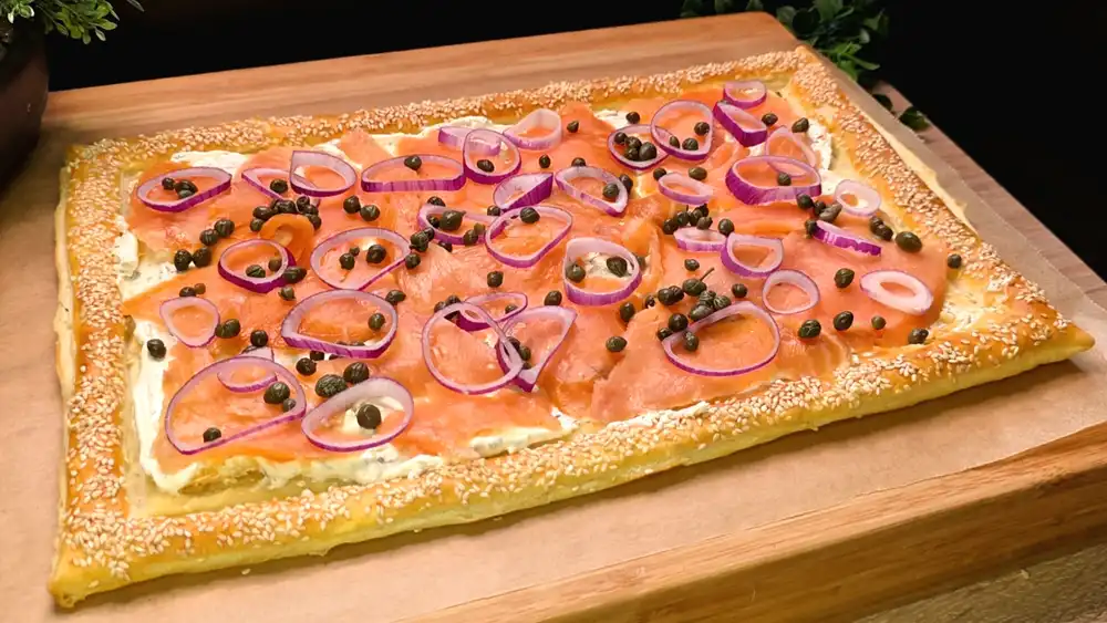 Goldbraun gebackene Blätterteig-Lachs-Tarte mit Kräuterfrischkäse, Räucherlachs, roten Zwiebelringen und Kapern, veredelt mit einem Sesamrand.