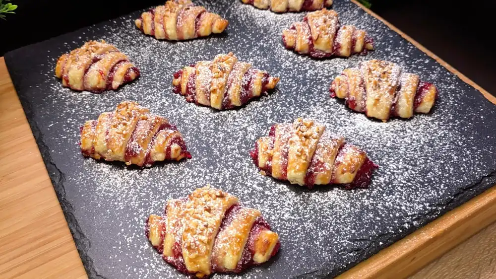 Goldbraun gebackene Blätterteig-Croissants mit Marmeladenfüllung und Puderzucker auf einem Teller.
