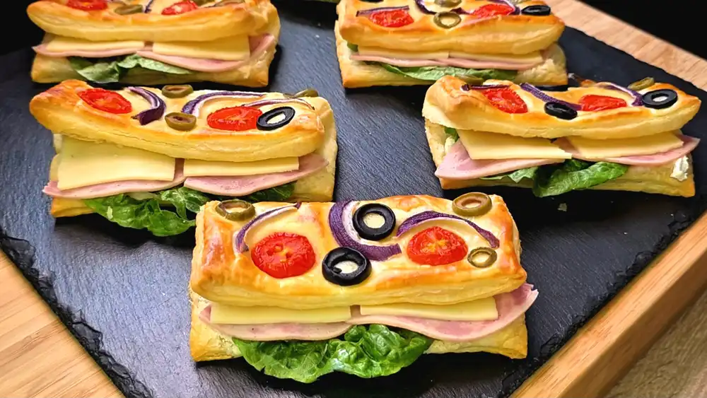 Goldbraun gebackene, aufgeschnittene Blätterteig-Sandwichbrötchen, belegt mit knackigem Salat, saftigem Schinken, Käse und cremigem Frischkäse, auf der Oberseite mit Tomaten, roten Zwiebeln und Oliven gebacken.