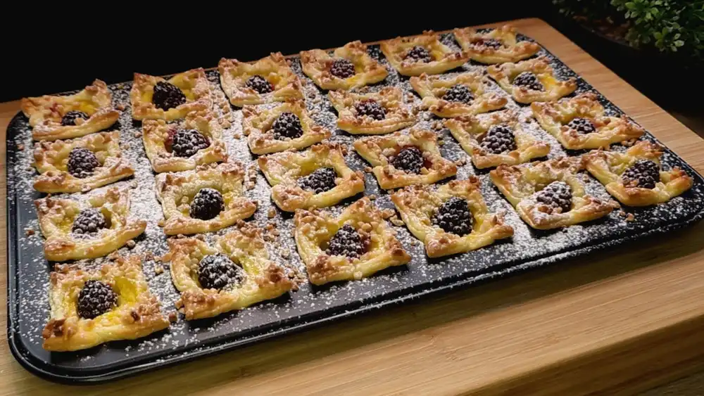 Eine Etagere voll mit 24 frisch gebackenen, krossen Blätterteig-Mini-Muffins, gefüllt mit Vanillepudding und je einer saftigen Brombeere, bestäubt mit Puderzucker.