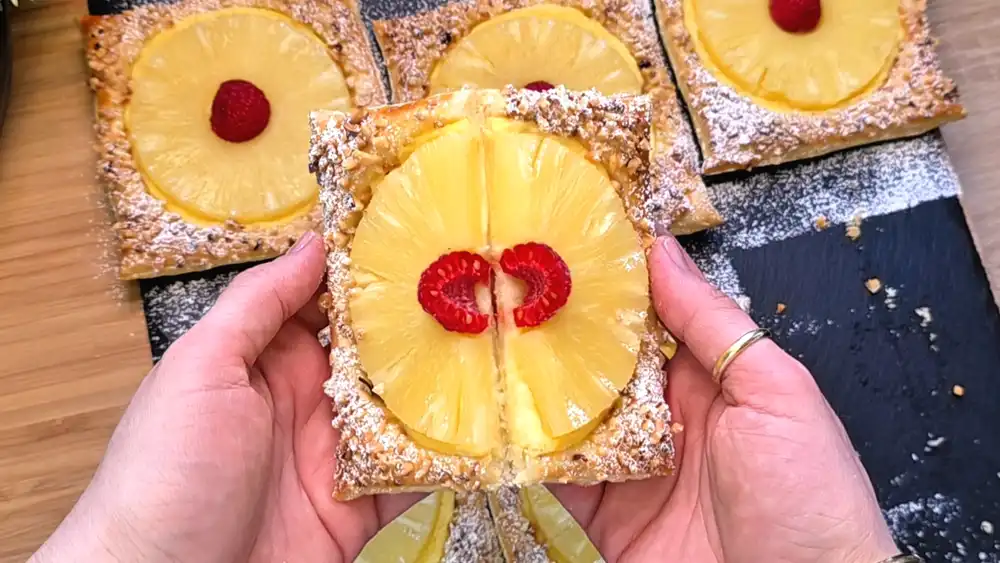 Detailansicht einer aufgeschnittenen Blätterteig-Schnitte mit einer saftig-gelben Ananasscheibe auf dickem Vanillepudding und gerösteten Nüssen.