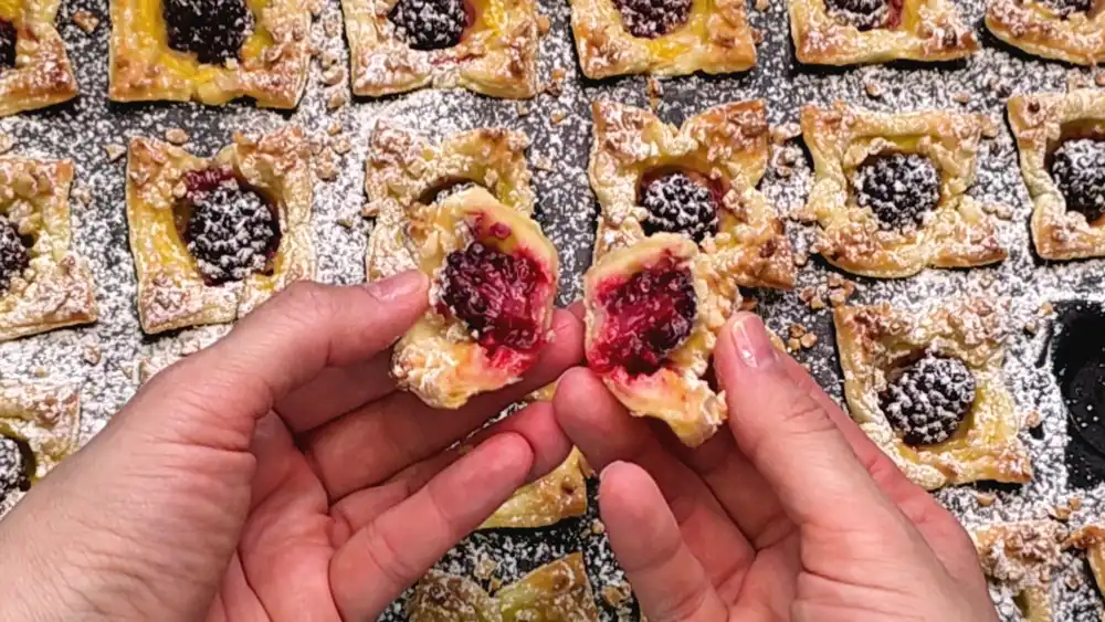 Detailansicht eines angebissenen Blätterteig-Muffins, die den cremigen Vanillepudding, die fruchtige Brombeere und die gerösteten Mandeln auf der krossen Kruste zeigt.