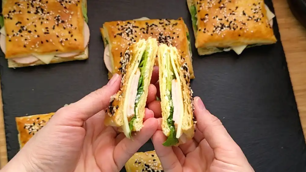 Detailansicht eines einzelnen, golden gebackenen Blätterteig-Sandwichs, das dick mit Sesam bestreut ist und bei dem die frischen Salatblätter und Schinkenscheiben saftig herausschauen.