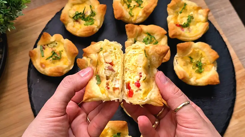 Detailansicht eines frisch gebackenen Blätterteig-Eiermuffins, bei dem die fluffige Eifüllung, rote Paprikastücke und die goldgelbe, knusprige Käsekruste perfekt zu sehen sind.