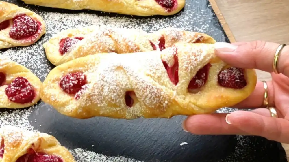 Detailansicht einer gebackenen Blätterteigtasche mit sichtbarer Quarkfüllung, heißen Himbeeren und Puderzucker.