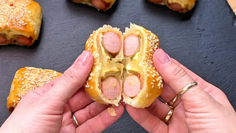 Detailansicht eines frisch gebackenen Hot-Dog-Häppchens mit sichtbarem Würstchen, Gewürzgurkenscheibe, Ketchup und Senf.