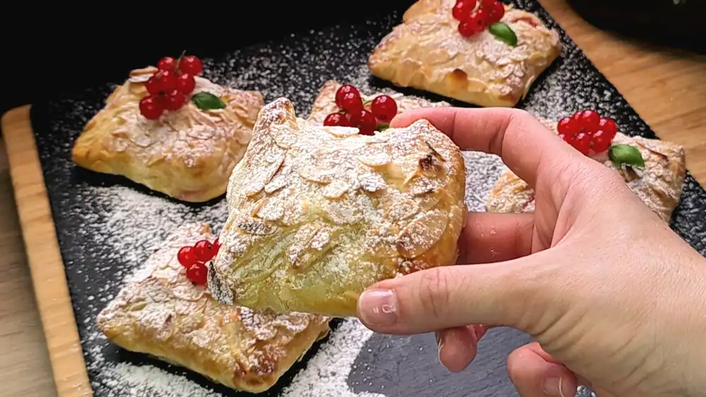 Detailansicht einer aufgeschnittenen Blätterteigtasche, bei der die weiße Quarkcreme und die prallen, roten Johannisbeeren unter den krossen Teigfalten hervorheben.