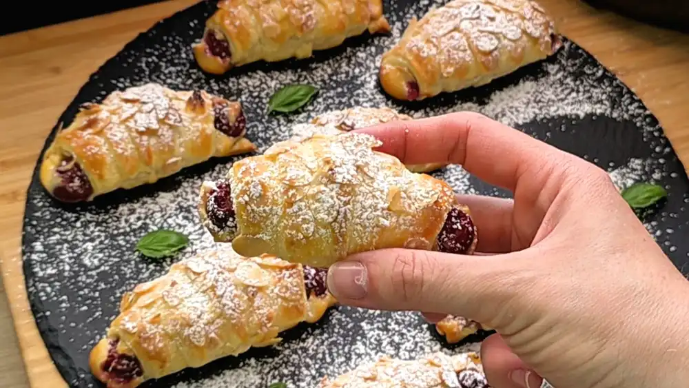 Detailansicht eines frisch gebackenen Quark-Croissants mit krossen Mandelblättchen, Puderzucker und sichtbarer Kirschfüllung.