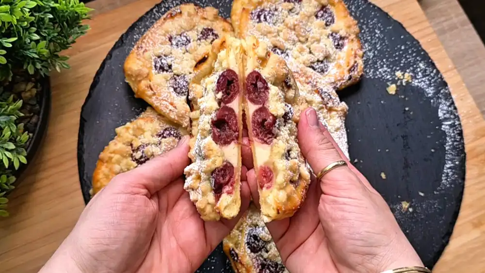 Detailansicht eines frisch gebackenen Mini-Quark-Kirsch-Kuchens aus Blätterteig, bei dem die cremige Quarkfüllung und die dicken Knusperstreusel unter dem Puderzucker glänzen.