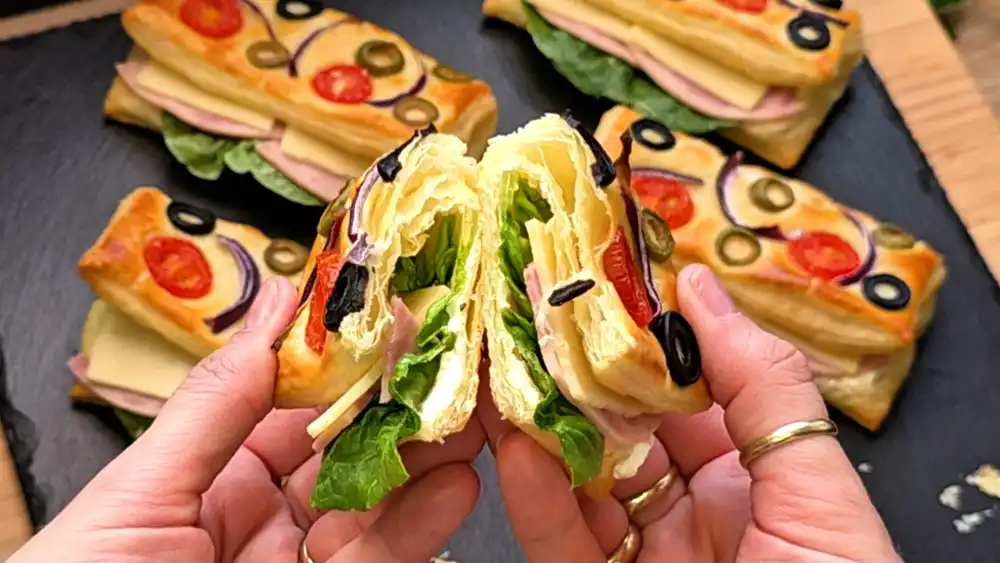Detailansicht der krossen Kruste eines Blätterteig-Brötchens mit eingebackenen Cocktailtomaten, roten Zwiebelringen und schwarzen Oliven.