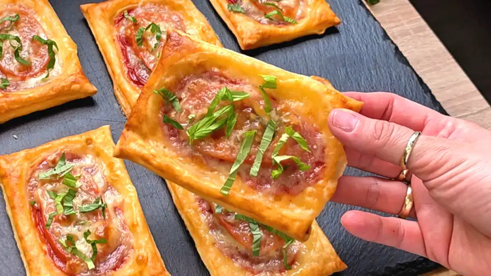 Detailansicht eines frisch gebackenen Pizza-Häppchens mit sichtbarer Salami, Bläschen werfendem Mozzarella und frischem Basilikumblatt.