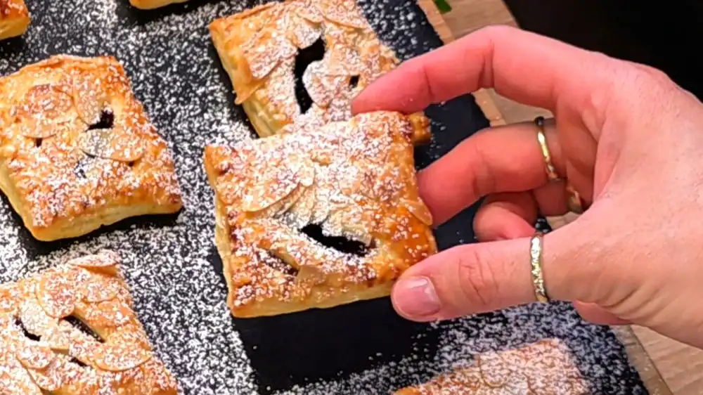 Detailansicht einer aufgeschnittenen Blätterteigtasche, bei der die gelbe Vanille-Quarkcreme und die dunkle Pflaumenmarmelade unter den krossen Teigstreifen hervorblitzen.