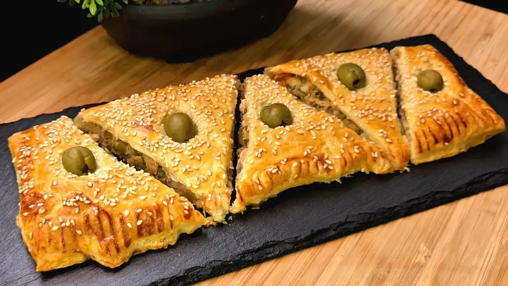 Goldbraun gebackene Thunfisch-Dreiecke aus Blätterteig mit Sesamsamen und ganzen grünen Oliven garniert.