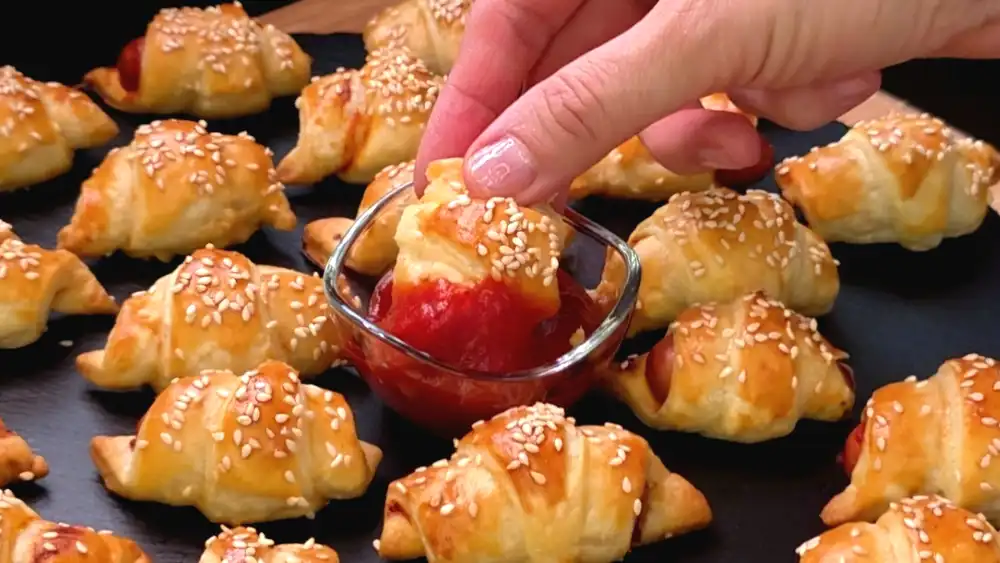 Detailansicht eines frisch gebackenen Würstchens im Schlafrock mit Sesamkruste und Tomatensoßen-Dip.