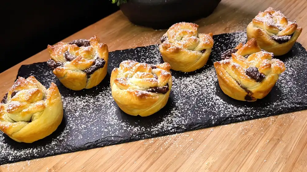 Eine Servierplatte mit sieben goldbraun gebackenen, krossen Blätterteig-Nutella-Rosen aus der Muffinform, fein mit Puderzucker bestäubt.