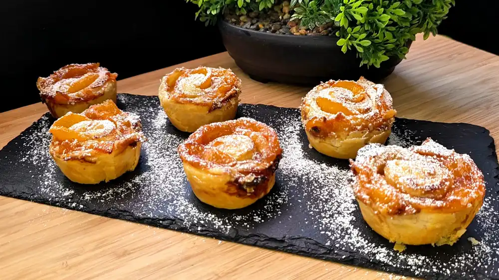 Goldbraun gebackene Blätterteig-Rosen mit fächerförmig eingewickelten Pfirsichscheiben in einer Muffinform, fein bestäubt mit Puderzucker.