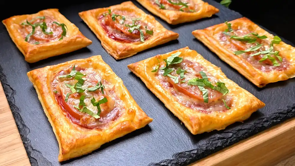 Goldbraun gebackene Mini-Pizzastücke aus Blätterteig mit Salami, geschmolzenem Mozzarella, einer Tomatenscheibe und frischem Basilikum.