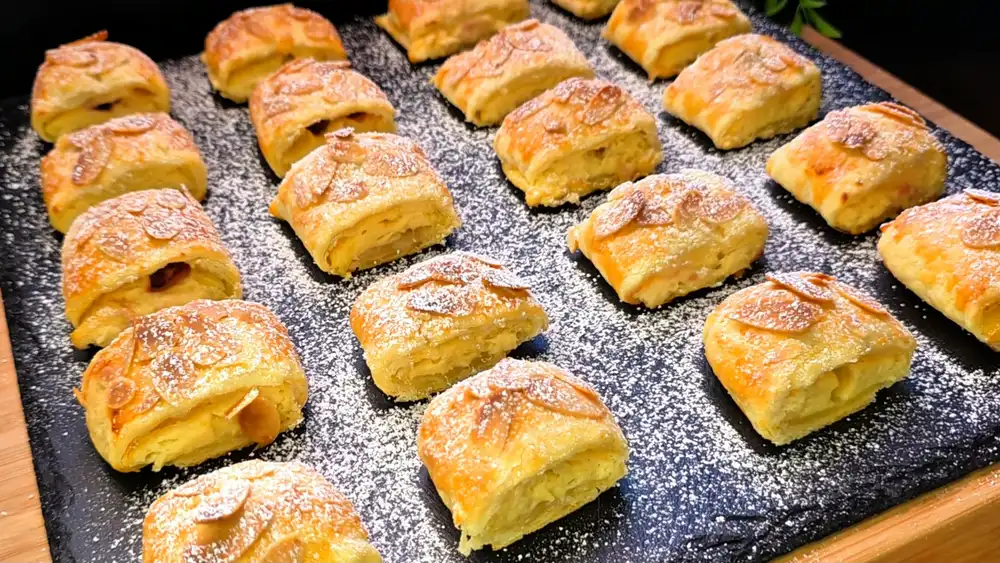 Eine Platte voller goldbraun gebackener Blätterteig-Quark-Kirsch-Häppchen mit feinen Einschnitten, Mandelkruste und Puderzucker-Dekoration im Garten.